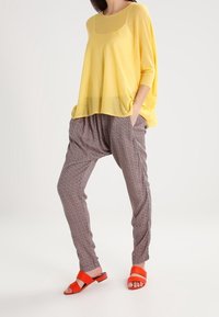 Gelbes, übergroßes Stricktop, gemusterte graue Haremshosen und orangefarbene flache Sandalen. Leichter, atmungsaktiver Stoff mit Taschen und lockerer Passform.