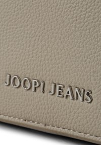 Beige nahkakassi, jossa on kohokuvioitu "JOOP! JEANS" -logo hopeana ja hienovaraiset ompeluyksityiskohdat reunoilla.