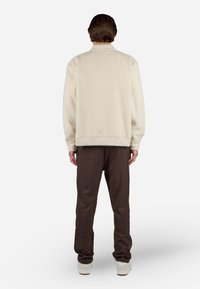 Cream-colored fleece jacket with a textured finish, paired with dark brown pants; view from the back highlighting the relaxed fit and movement.
