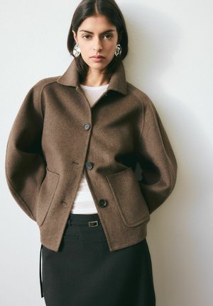 Woman wearing brown buttoned wool jacket over white top and black skirt, standing against white wall, hands in jacket pockets.
