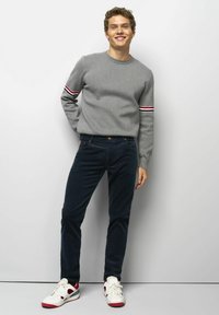 Grau gestrickter Pullover mit gestreiften Akzenten, dunkelblaue Hose und weiße Sneakers mit roten Details. Das Modell steht mit entspannter Haltung vor einem hellen Hintergrund.