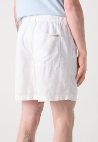 Valkoiset puuvillashortsit, joissa on joustava vyötärö, taka-tasku ja Calvin Klein -logotagi. Materiaali näyttää kevyeltä ja sileältä.