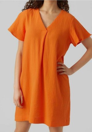 Femme portant une robe orange ample et texturée à manches courtes, col en V et pli frontal, debout avec une main sur la hanche.