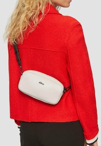 Weißer rechteckiger Crossbody-Beutel mit schwarzem Riemen, der eine glatte Textur und ein dezentes Logo aufweist, getragen über einer roten Jacke.