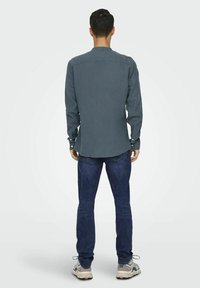 Camisa azul oscuro de botones con mangas largas, hecha de tela suave. La parte trasera presenta un canesú y un dobladillo curvado, combinada con jeans oscuros y zapatillas.