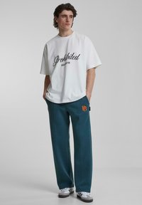 Hvit t-skjorte med korte ermer og svart kursiv «Prohibited Berlin New York» tekst, sammen med teal joggebukser med oransje logo.