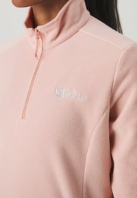 Ljusrosa fleece-tröja med kvartslång dragkedja och hög krage samt broderad logotyp. Mjuk textur med minimala sömdetaljer.