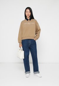 Brun genser med hvit logo, blå skjorte under, løse blå jeans, hvite sneakers og en liten hvit håndveske.