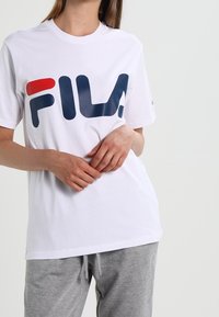 White cotton T-shirt featuring a large multicolored "FIL" logo in navy and red, paired with gray sweatpants. Simple, casual design.