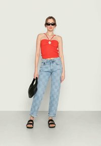 Top curto vermelho com alças finas e recorte em forma de chave na parte frontal, combinado com jeans azul claro xadrez, sandálias de plataforma pretas e uma bolsa preta.