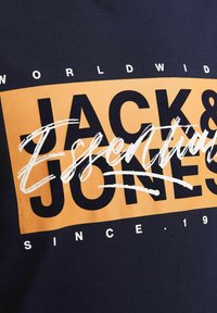 Námorně modré bavlněné tričko s velkým oranžovým obdélníkovým grafickým vzorem, na kterém je v tučném černém písmu nápis "JACK & JONES" a "ESSENTIALS" v bílém kurzívním písmu.