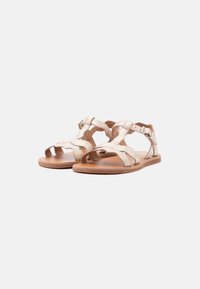 Μπεζ επίπεδες σανδάλια με υφασμένες λουριά, ανοιχτά δάχτυλα και αγκράφες στον αστράγαλο σε λευκό φόντο.