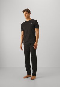 Fekete pamut póló rövid ujjakkal és egy kis fehér logóval a mellkasán, párosítva fekete, slim fit nadrággal, a modell mezítláb van.