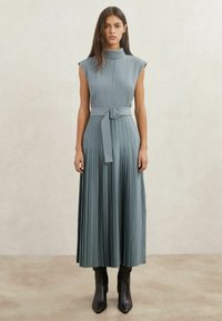 Plissé midi-jurk van zachtblauwe stof met een hoge kraag, een taille met ceintuur en een mouwloos ontwerp. Afgewerkt met zwarte laarzen met hak.