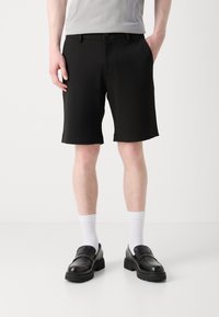 Shorts noirs en tissu lisse, avec une fermeture à bouton et des poches latérales en biais. Portés avec des chaussettes blanches et des mocassins noirs.