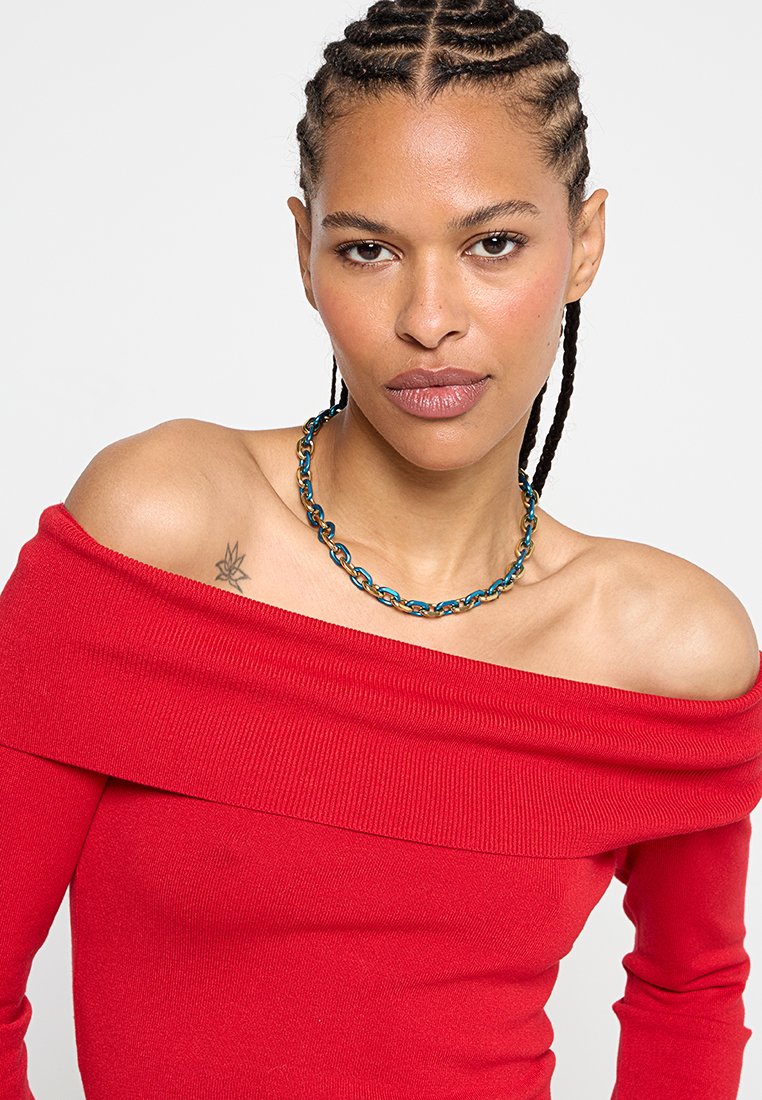 Red off-the-shoulder top with ribbed texture, paired with a multicolored chain necklace featuring blue and gold links.