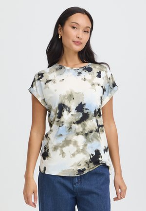 Femme aux longs cheveux foncés portant une chemise à manches courtes à motif abstrait avec des tons bleus, noirs et beiges et un jean bleu.