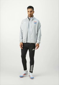 Graue Zip-Jacke mit Logo, schwarze enganliegende Hose mit weißen Streifen und farbenfrohe Sportschuhe mit blauen und roten Akzenten.