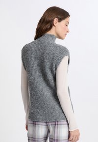 Maglione a collo alto a maniche corte in morbida maglia grigia, con finitura testurizzata. Indossato sopra una leggera maglietta a maniche lunghe e pantaloni a scacchi.