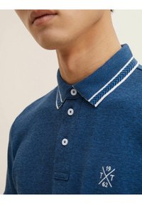 Lyhythihainen sininen poolopaita, jossa on tekstuuri kangas. Paidassa on kontrastivärinen kaulus kalanruotokuvioilla ja rintapielessä kirjailtu logo.