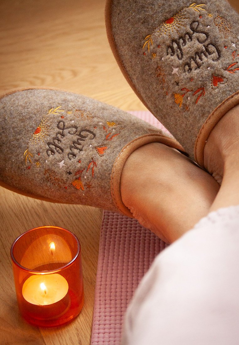 Gray felt slippers with embroidered floral patterns and text "Gute Seele." Next to a lit orange candle on a textured pink mat.