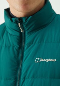 Nærbillede af en person iført en teal Berghaus zip-jakke med høj krave og synlig logo på brystet.