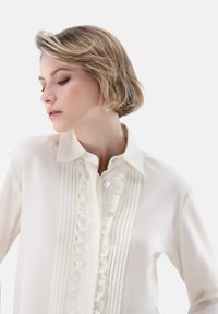 White blouse with ruffled front, pleated detailing, and a buttoned closure. Features a classic collar and smooth, soft fabric texture.