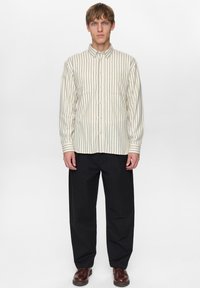 Overhemd in roomkleur met marineblauwe verticale strepen; voorzien van een kraag, lange mouwen en een borstzak. Gecombineerd met zwarte broek en bruine schoenen.