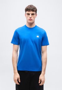 Νέος άνδρας που φοράει φωτεινό μπλε κοντομάνικο μπλουζάκι με μικρό λογότυπο στο στήθος, στέκεται μπροστά σε απλό ανοιχτό γκρι φόντο.