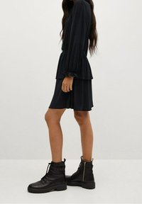 Vestido mini negro con capas y mangas largas, con puños fruncidos, combinado con botas de tobillo negras con cordones, que presentan un cierre lateral y una suela gruesa.
