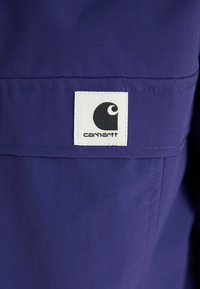 Tissu violet avec une texture lisse, présentant une étiquette blanche rectangulaire avec un logo noir et le texte "carhartt" au bas.
