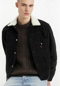 Young man wearing a black corduroy jacket with white shearling collar, dark cable-knit sweater, and light blue jeans against a white background.