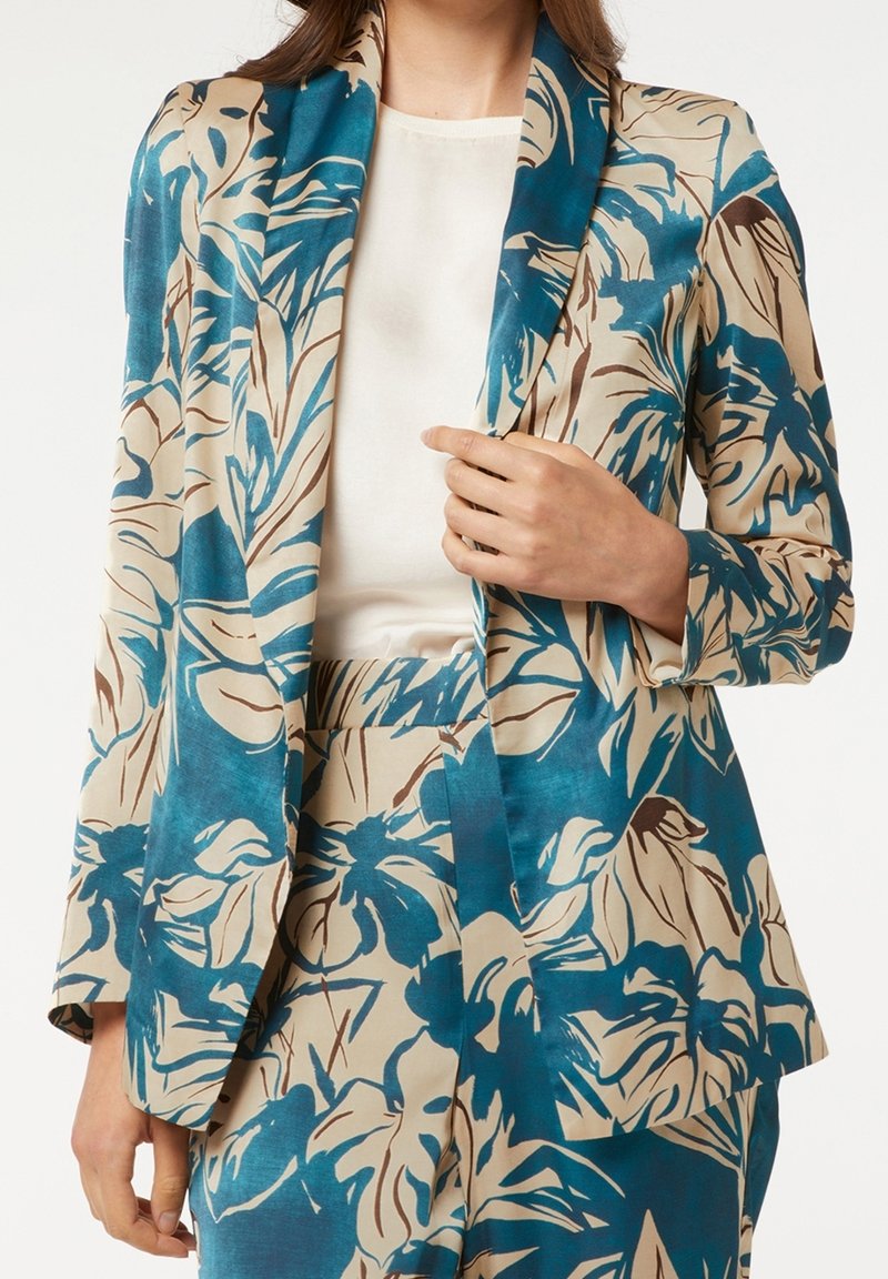Femme portant un blazer à imprimé floral bleu sarcelle et beige avec un pantalon assorti, associé à un haut blanc, tenant le revers du blazer.