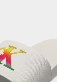 Λευκό σανδάλι με φαρδιά υφασμάτινη λωρίδα που φέρει πολύχρωμο διαβαθμισμένο λογότυπο "CK" στην κορυφή.