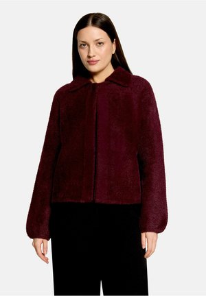 Woman with straight dark hair wears a dark red fuzzy jacket over black pants, standing against a plain light background.