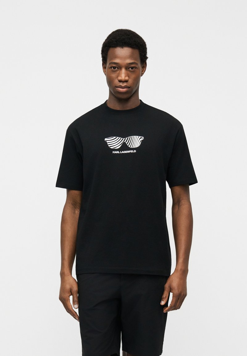 Νεαρός άνδρας με μαύρο t-shirt με γραφικό λευκά ριγέ γυαλιά ηλίου και το κείμενο "KARL LAGERFELD", στέκεται μπροστά σε ένα απλό φωτεινό φόντο.