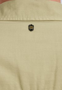 Beige shirt fabric with a smooth texture, featuring a black PME Legend tag secured to the back collar by a small black fastener.