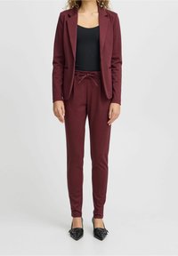 Burgundin takki, jossa on viistetyt kaulukset, yhdistettynä saman värisiin kapeneviin housuihin, joissa on nyöritetty vyötärö. Mustat kärjelliset balleriinat.