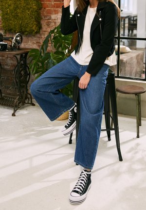 Person, der sidder på en sort metalstol, iført en sort jakke, hvid skjorte, brede blå jeans og sorte high-top sneakers indendørs ved siden af planter.