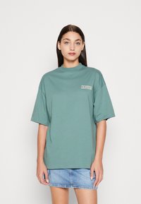Green cotton t-shirt with a high neckline and loose fit. Features a white "CALIFORNIA" print on the left side. Short sleeves and smooth texture.