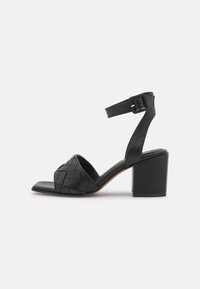 Schwarze Ledersandalen mit Absatz, die ein geflochtenes Design am Zehenriemen, einen Knöchelriemen mit Schnalle und einen quadratischen Blockabsatz aufweisen.