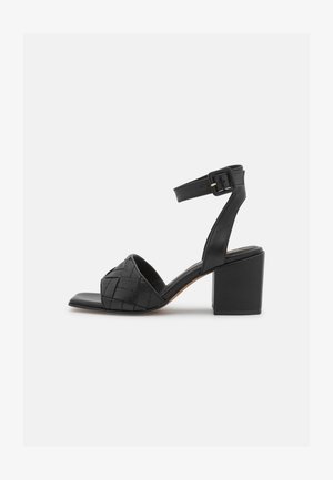Black leather heeled sandals featuring a woven design on the toe strap, an ankle strap with a buckle, and a square block heel.