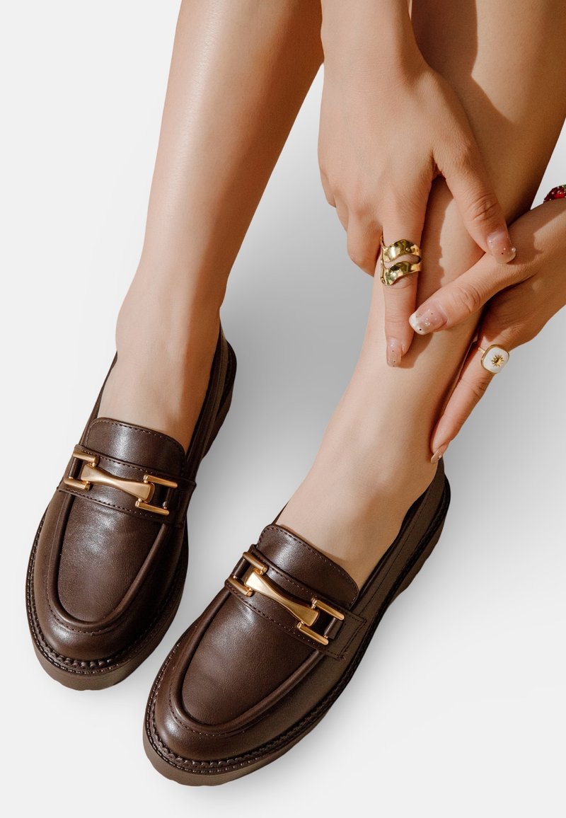 Mocassins en cuir marron avec des accents de quincaillerie dorée, présentant un embout arrondi et des détails de couture. Portés avec les mains reposant sur les jambes.