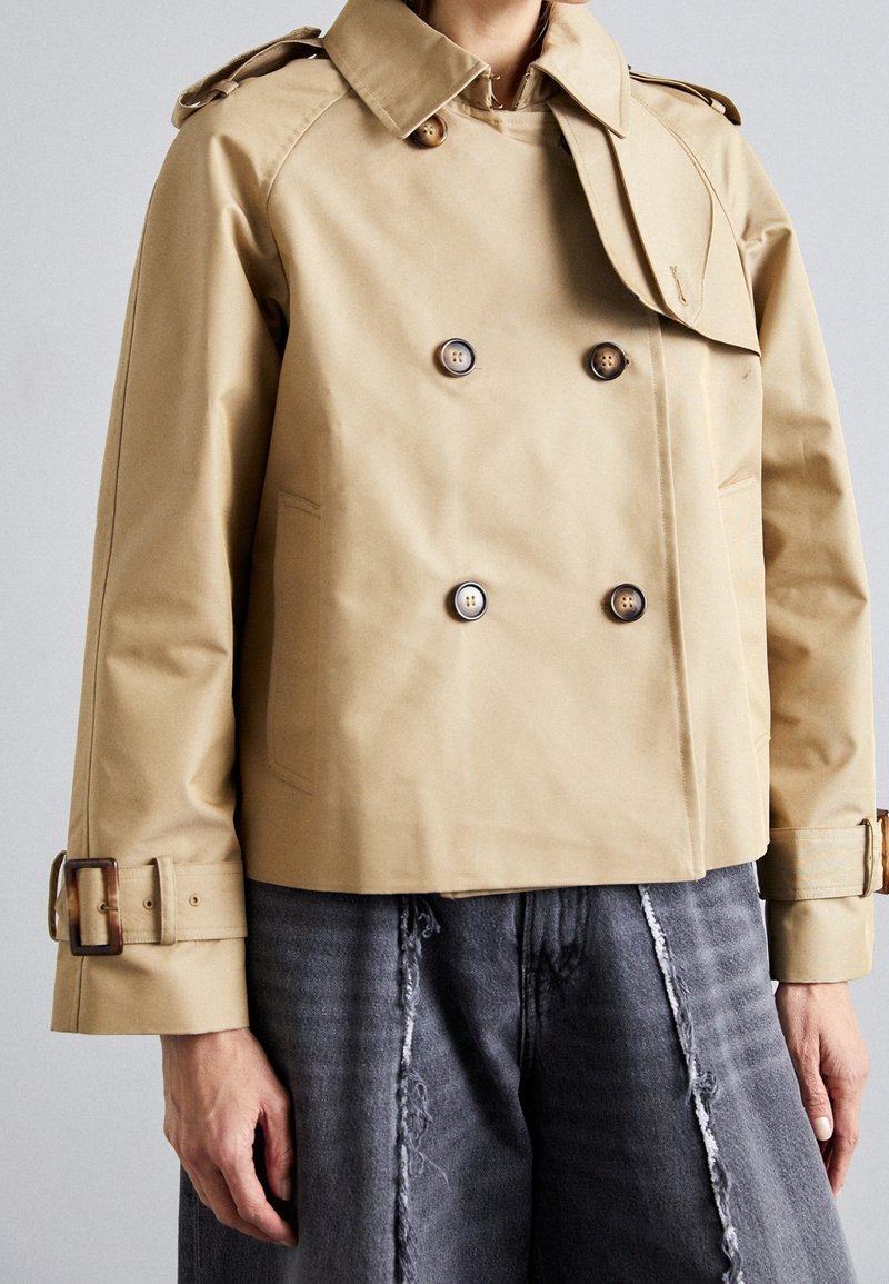 Personne portant un trench-coat beige court à double boutonnage avec épaulettes et poignets à boucles, associé à un jean gris foncé effiloché.