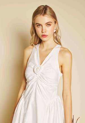 Tânără cu păr blond, purtând o rochie albă fără mâneci, cu detaliu floral tip rozetă și cercei în formă de inimă, pe un fundal bej.