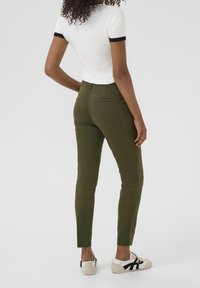 Pantaloni aderenti verde oliva con una texture liscia, dotati di due tasche posteriori e un design a gamba tapered, abbinati a sneakers casual.