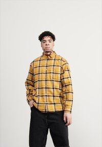 Camisa de franela a cuadros amarillos con cierre de botones, mangas largas y corte relajado. Jeans oscuros, sin patrones o adornos visibles.