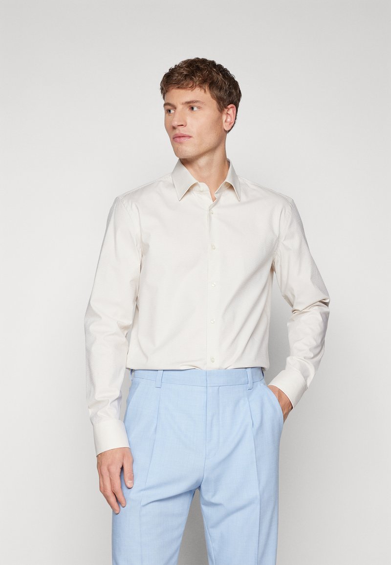 Maglietta a maniche lunghe color beige chiaro con chiusura a bottoni e una sottile texture, abbinata a pantaloni tagliati in blu chiaro, che mostrano una silhouette aderente.