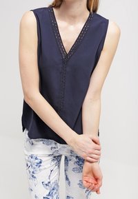 Marinblå ärmlös topp med V-ringning och spetsdetaljer, mjukt tyg och avslappnad passform, tillsammans med vita byxor med blommönster.