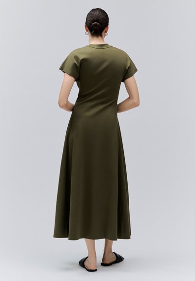 Femme debout de dos portant une robe midi à manches courtes vert olive et des sandales noires sans lacets sur un fond uni.